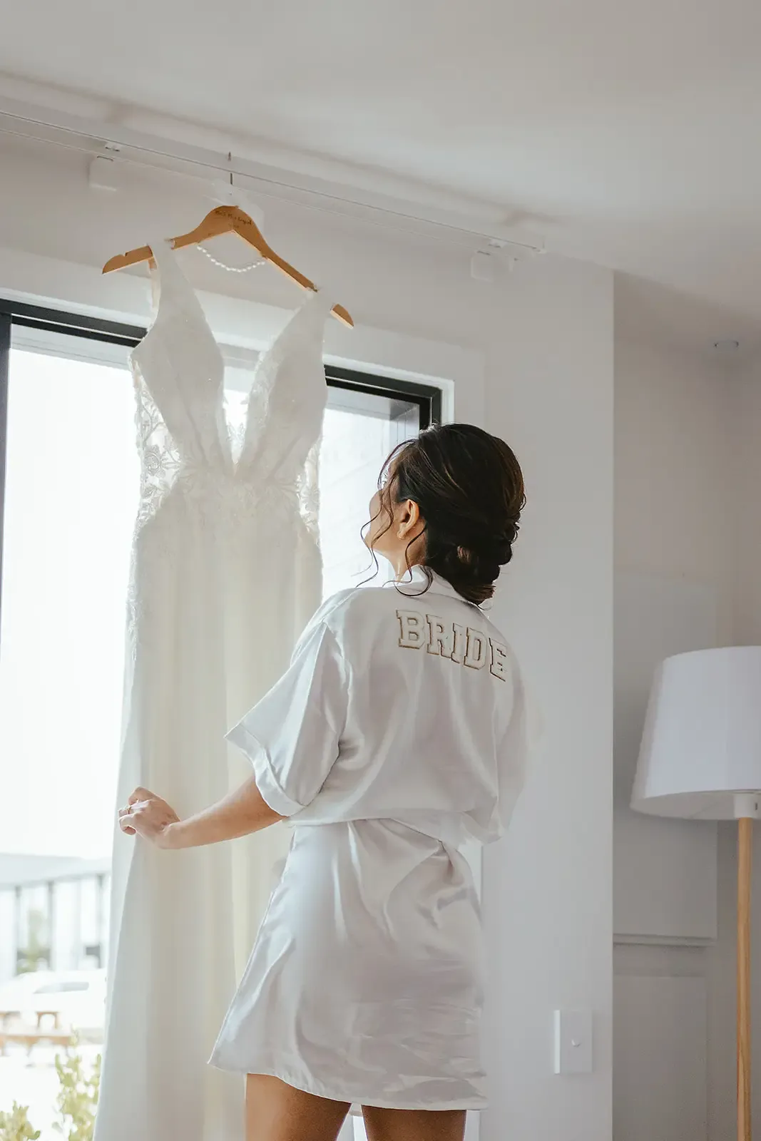 Bride in a robe beside her wedding dress hanging by a window