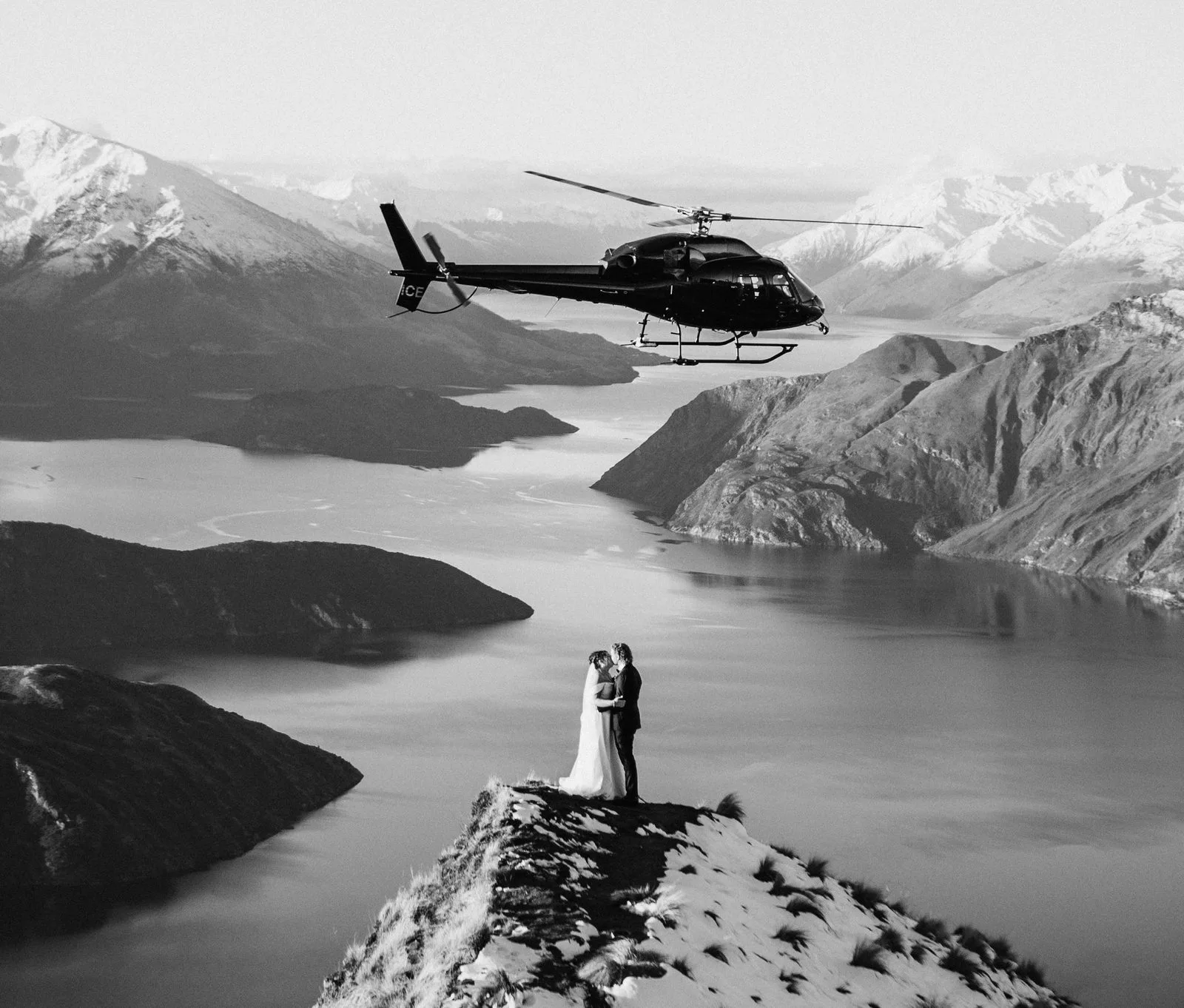 Black and white frame of a couple on the Coromandel Peak ridge with a helicopter above Lake Wānaka