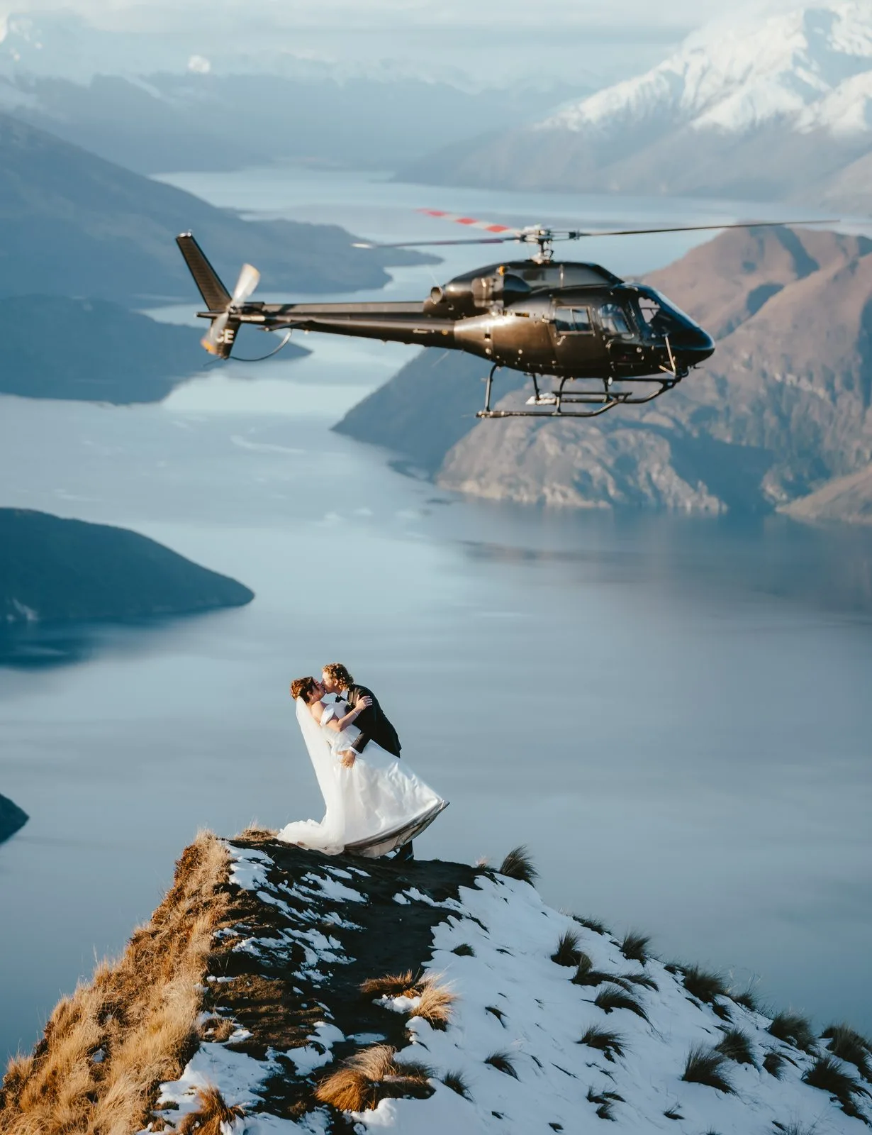Bride and groom kissing on the Coromandel Peak ridge in winter as a helicopter passes above Lake Wānaka