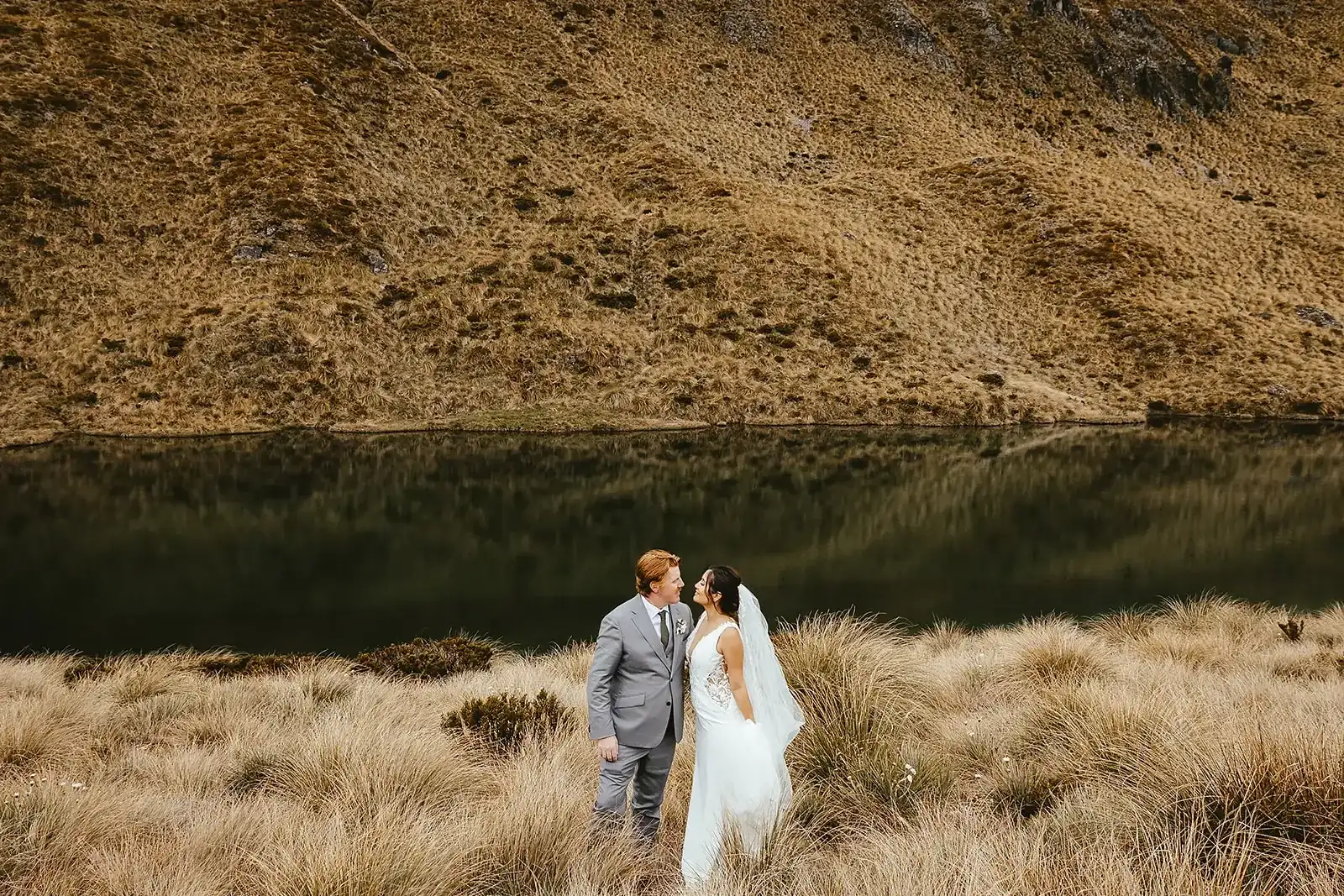 Couple standing in tussock beside an alpine tarn reflection