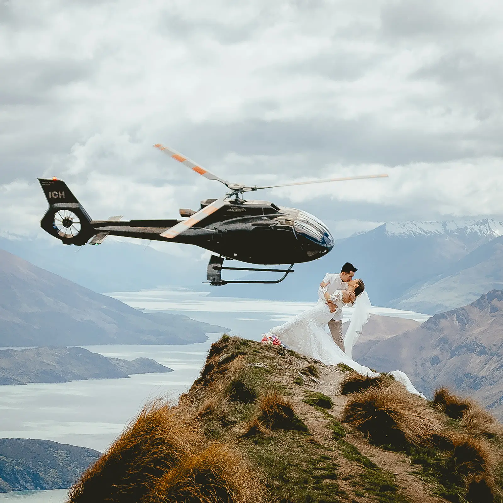 Couple in a dip kiss at Coromandel Peak beside the helicopter, Lake Wānaka behind