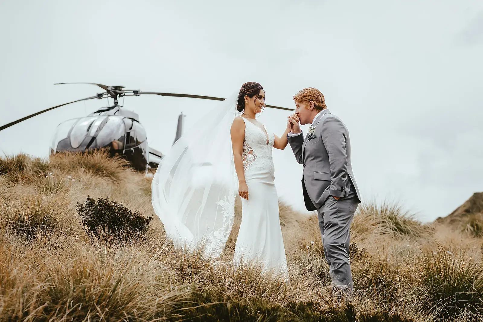 Groom kissing the bride's hand beside a helicopter, veil flowing