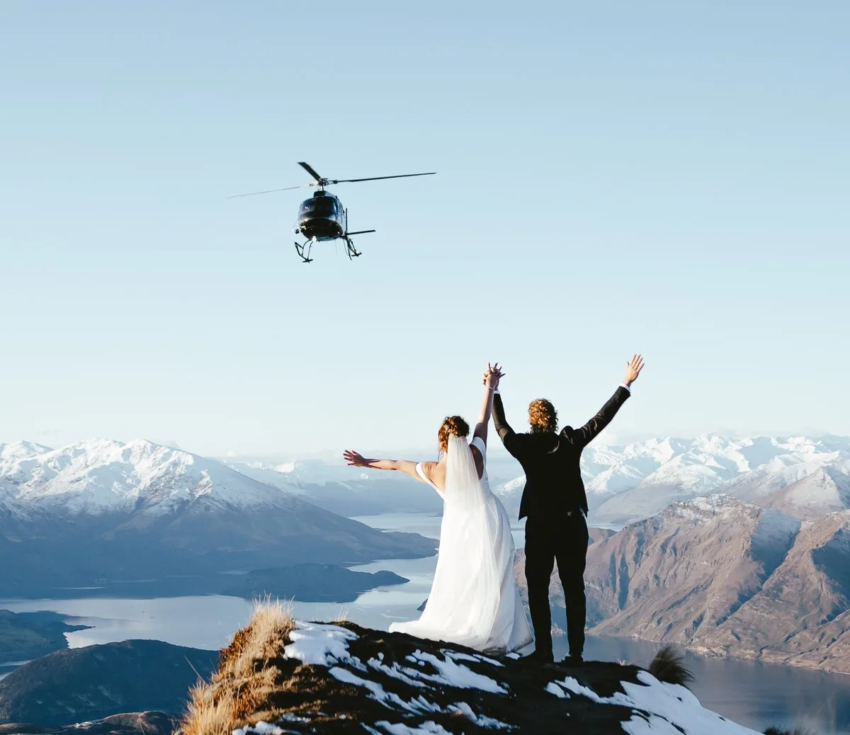 Couple with arms raised on a snowy ridge above Lake Wānaka as a helicopter flies past