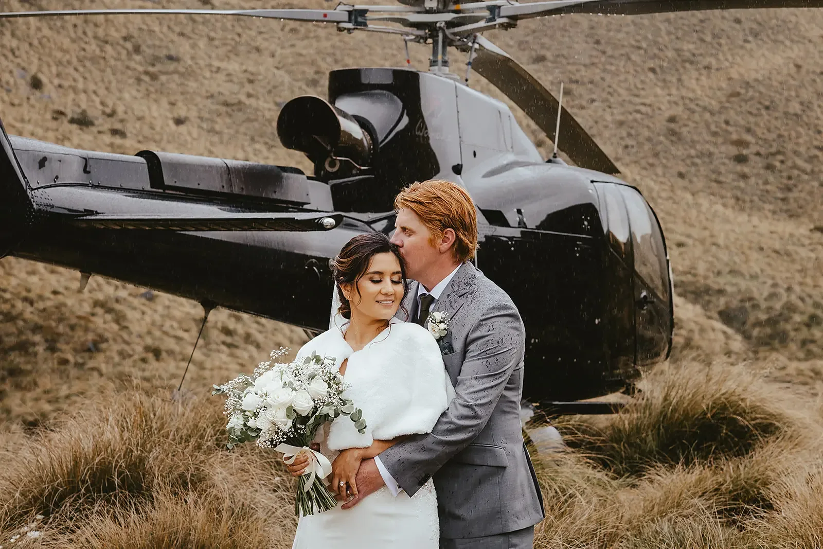 Groom kissing the bride's forehead beside a helicopter