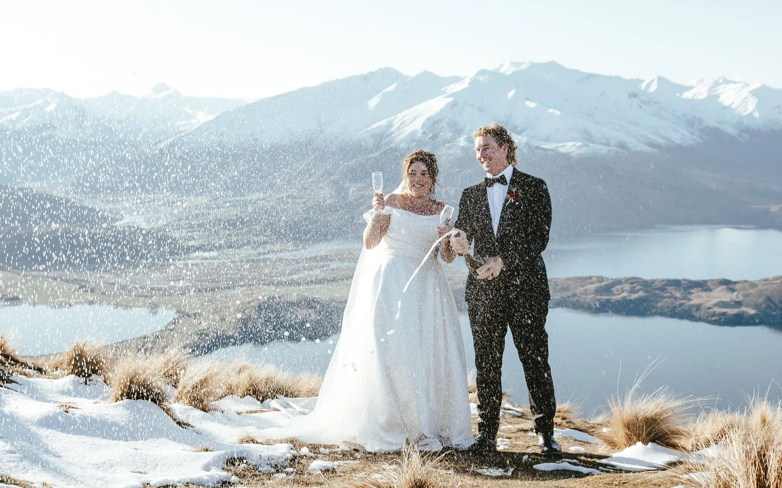 Couple raising champagne glasses on a snowy ridge at Coromandel Peak with Lake Wānaka behind them
